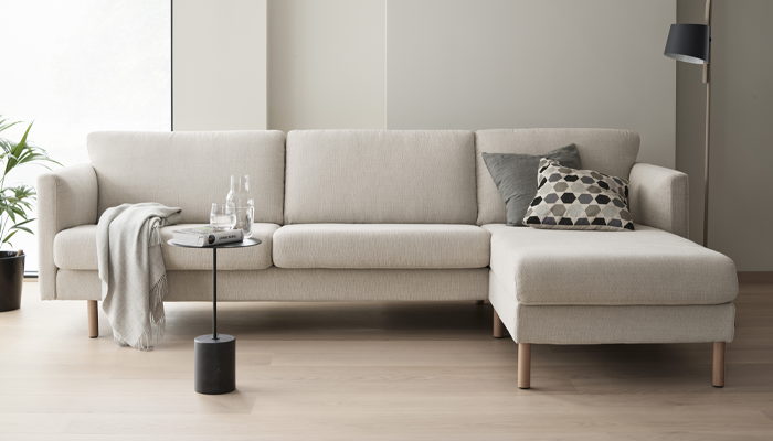 Stressless Sofa • Cremefarbenes Ecksofa vor einer weißen Wand, dekoriert mit Kissen und einer Decke. Davor ein kleiner Tisch mit Gläsern • Meyerhoff