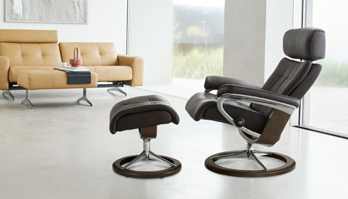 Stressless Sessel • Brauner Sessel mit Hocker vor einer Fensterfront. Im Hintergrund ein helbraunes Sofa • Meyerhoff Stressless Sessel • Brauner Sessel mit Hocker vor einer Fensterfront. Im Hintergrund ein helbraunes Sofa • Meyerhoff